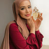 A woman wearing a dusty rose chiffon hijab scarf and a maroon top, holding a glass perfume bottle.