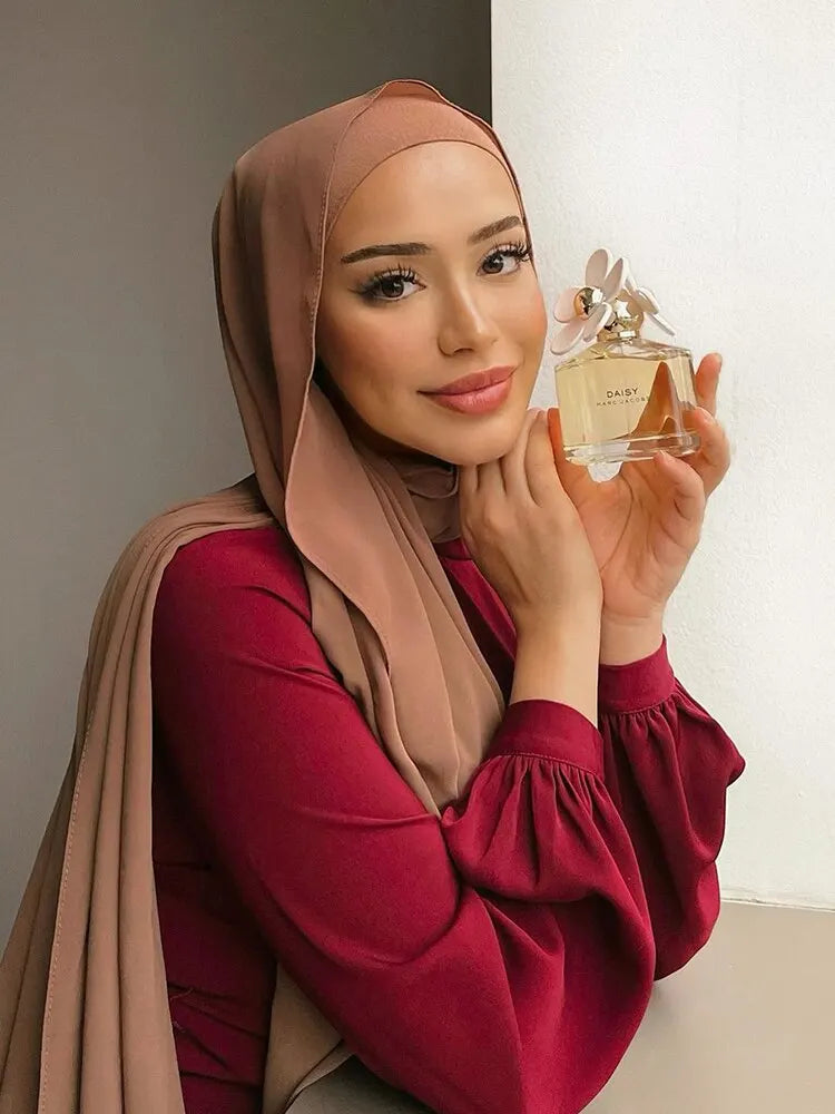 A woman wearing a dusty rose chiffon hijab scarf and a maroon top, holding a glass perfume bottle.