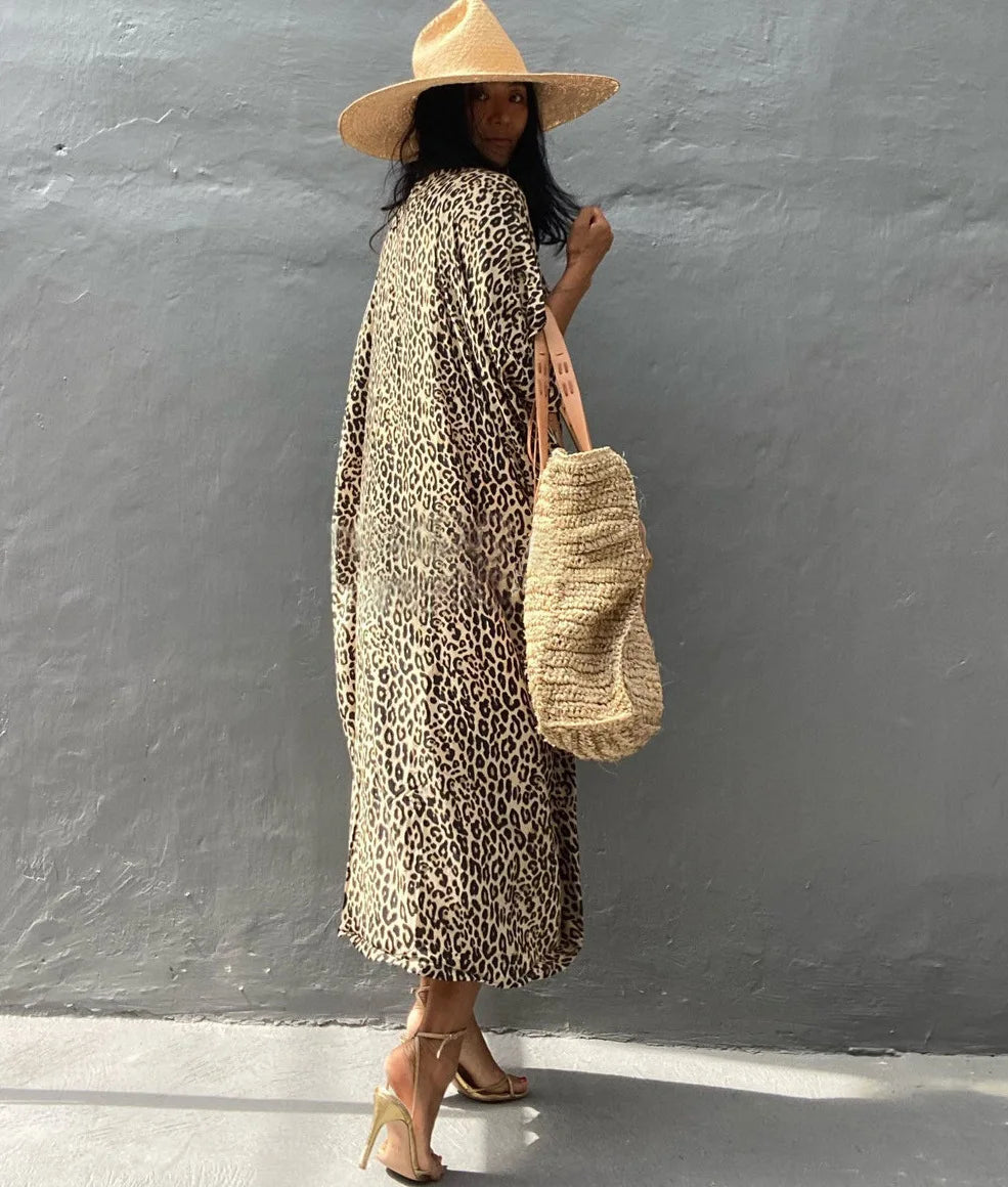 Eine Frau in einem Leopardenmuster-Maxikleid und einem Strohhut vor einer grauen Wand.