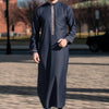 Un homme portant une robe musulmane bleu foncé avec broderie rétro verticale, debout à l'extérieur.
