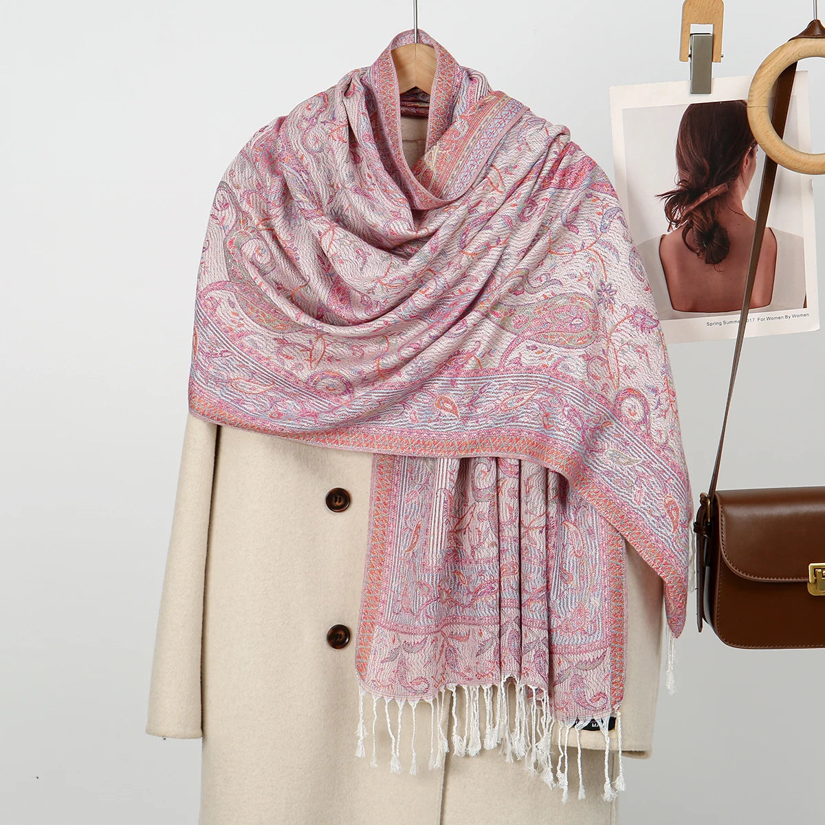 Chal de bordado paisley rosa y blanco sobre un abrigo beige, con un bolso de cuero marrón cercano.