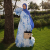 Eine Frau in einem eleganten blau-weißen Blumenmuster-Abaya und Kaftan steht in einem üppigen grünen Garten.