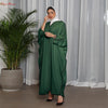Una mujer vistiendo una abaya verde esmeralda oversize con ribete negro y un hijab blanco, de pie frente a una cortina transp