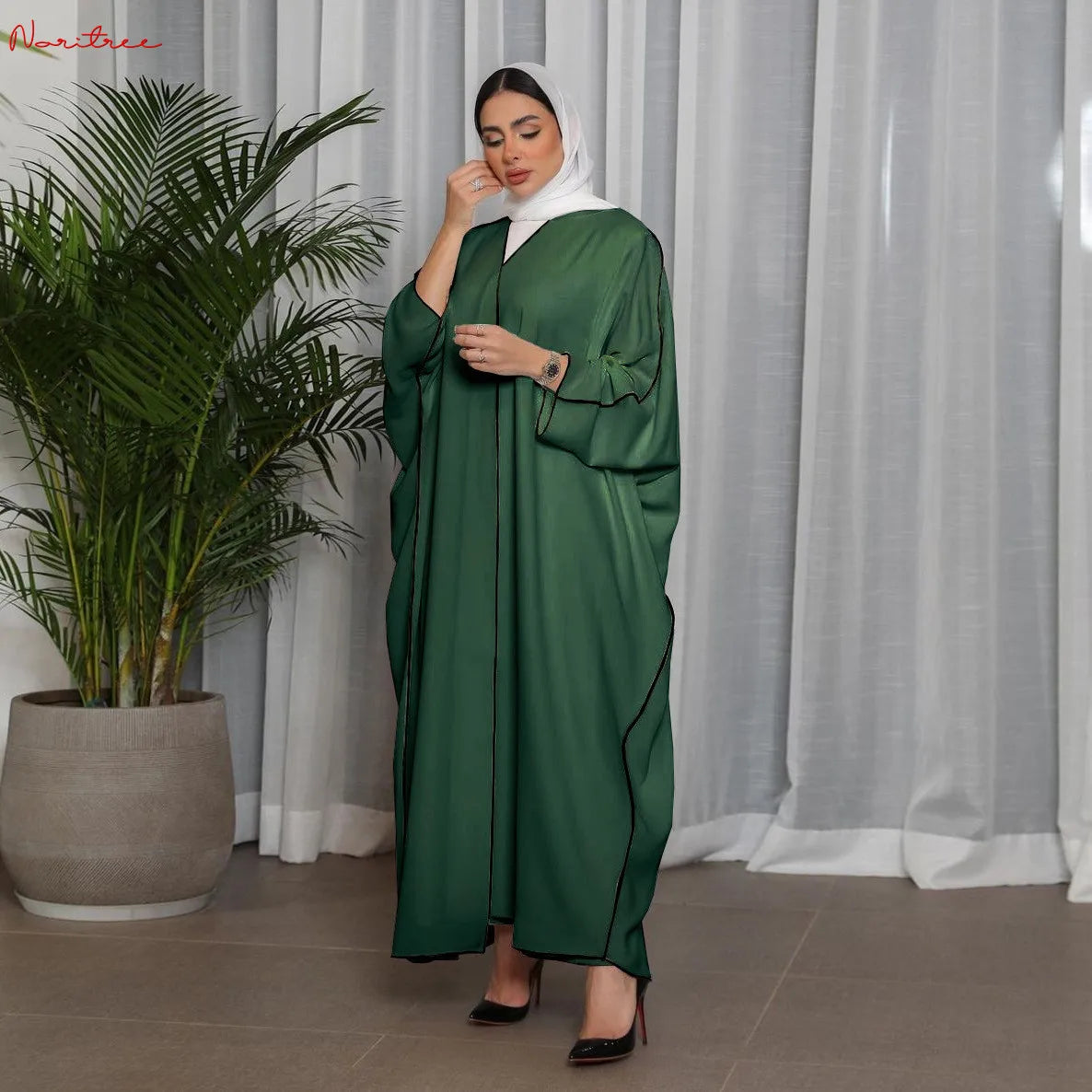 Una mujer vistiendo una abaya verde esmeralda oversize con ribete negro y un hijab blanco, de pie frente a una cortina transp