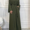 A woman wearing an olive green abaya and kaftan set, standing against a white paneled wall.