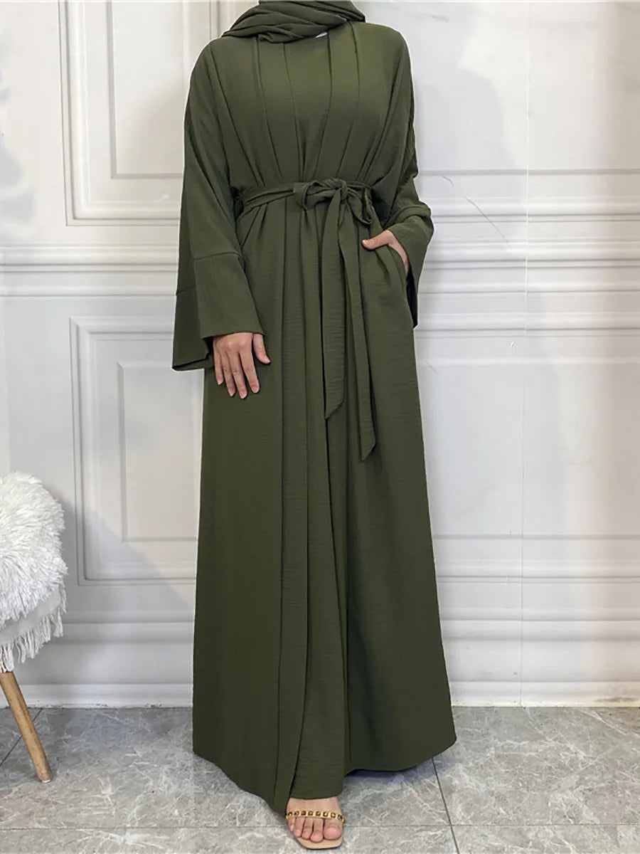 A woman wearing an olive green abaya and kaftan set, standing against a white paneled wall.