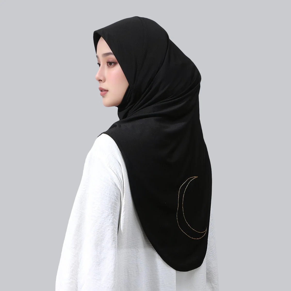 Une femme portant un hijab noir avec une broderie de croissant de lune doré sur un fond gris clair.