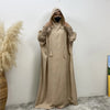 Un mannequin portant une abaya beige avec capuche et manches batwing, debout contre un mur blanc.