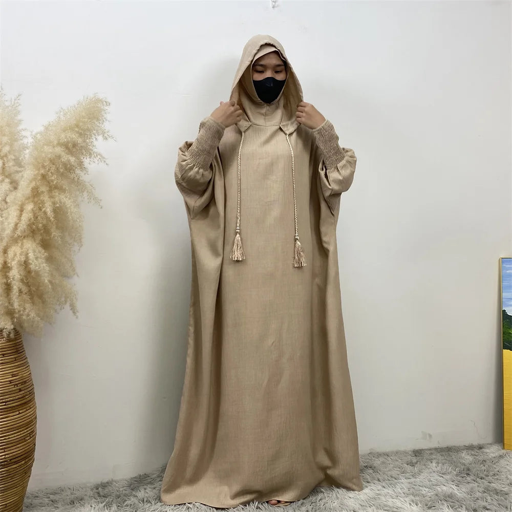 Un mannequin portant une abaya beige avec capuche et manches batwing, debout contre un mur blanc.