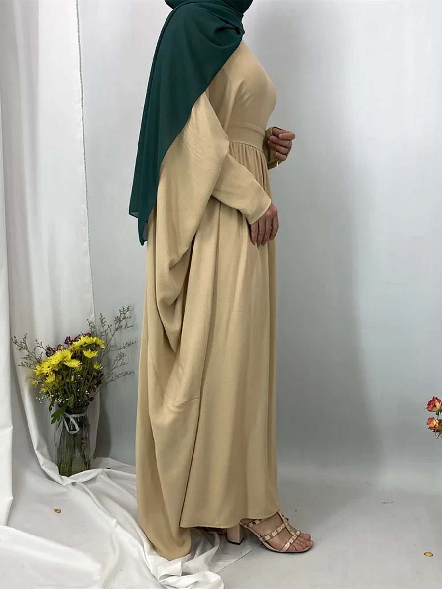 Eine Frau trägt eine elegante beige Batwing-Abaya und einen dunkelgrünen Hijab in einer hellen Umgebung.