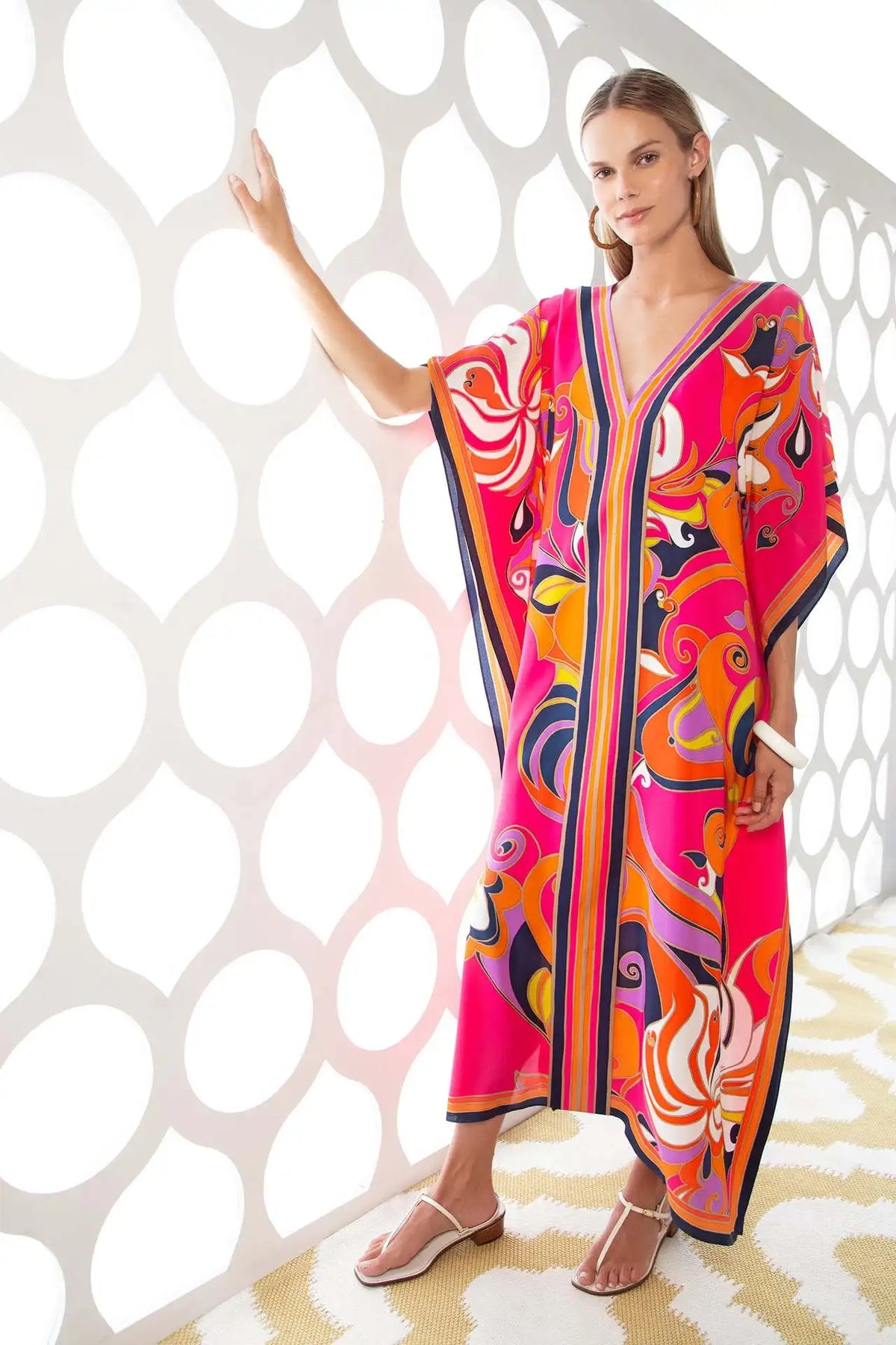 Modelo vistiendo un caftán de playa estampado bohemio rosa y naranja vibrante contra una pared blanca estampada.