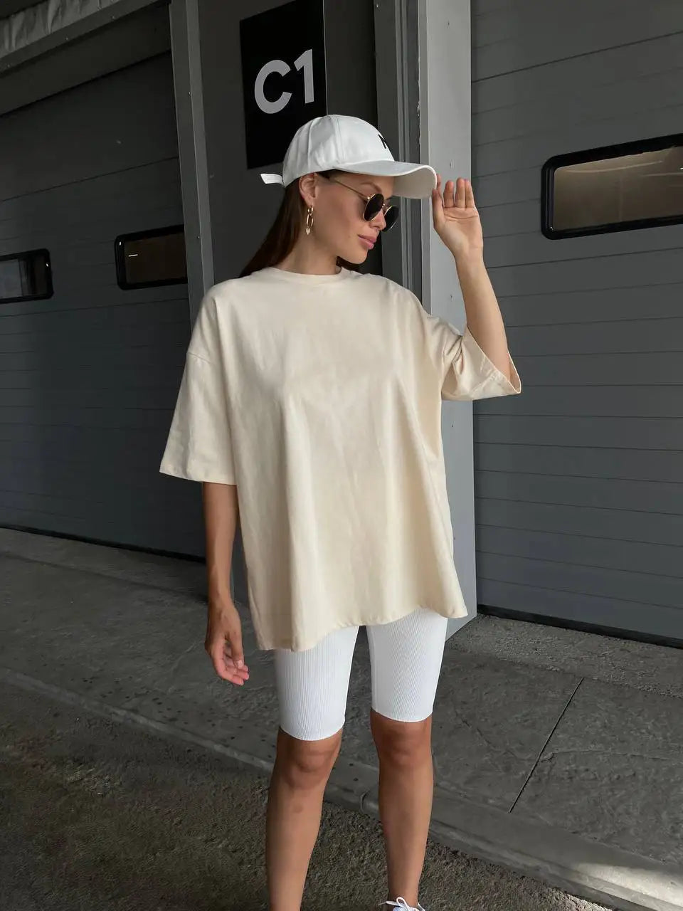 Una mujer viste una camiseta básica de algodón oversize color crema y pantalones blancos frente a una puerta de garaje gris.