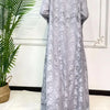 Une longue abaya en dentelle grise pour femmes, exposée contre un mur blanc à côté d'une plante verte.