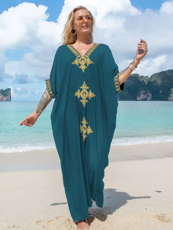 A woman wearing a teal kaftan dress with gold embroidery stands on a beach with tropical islands in the background.