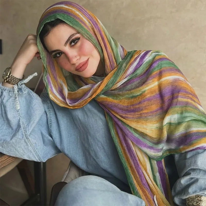 Una mujer vistiendo un pañuelo hijab a rayas multicolores y una camisa de mezclilla, mirando directamente a la cámara.