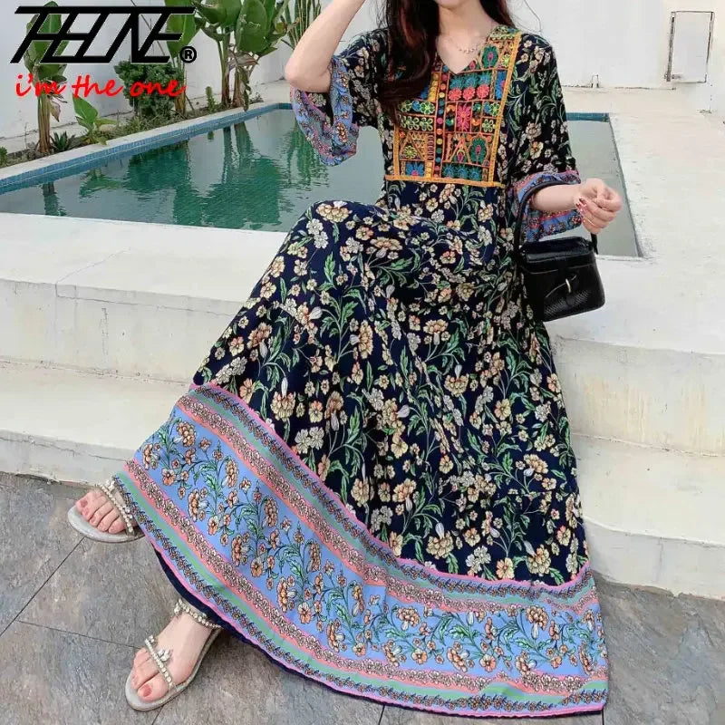 Eine Frau in einem dunkelblauen, floral bestickten Maxi-Kleid mit rosa Zierrand sitzt draußen am Pool.