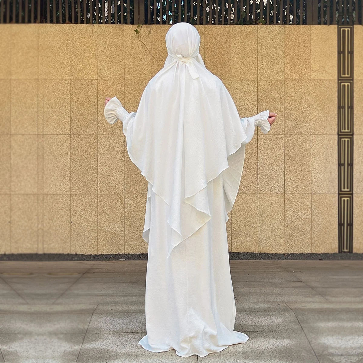 A full-length shot of a woman wearing an elegant white abaya and khimar set against a beige wall.