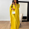 Una mujer vistiendo una abaya de satén amarillo brillante con forma de ala y un bolso blanco, posando frente a un gran espejo