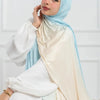 Una mujer vistiendo un pañuelo hijab de algodón con degradado azul claro y beige de doble cara sobre una blusa blanca.