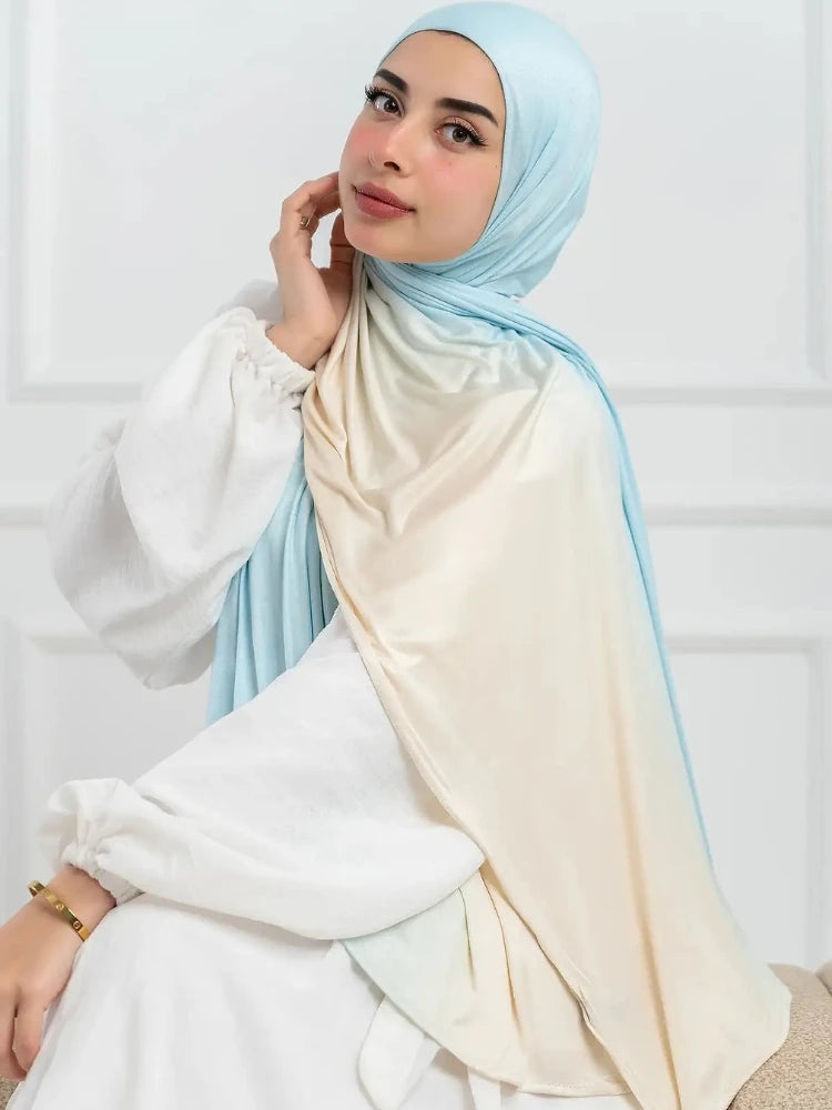 Una mujer vistiendo un pañuelo hijab de algodón con degradado azul claro y beige de doble cara sobre una blusa blanca.