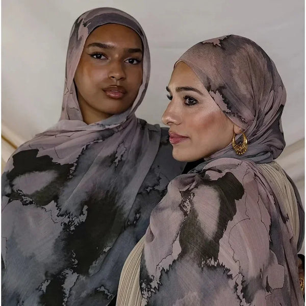 Two women wearing elegant printed voile hijab scarves in shades of gray and beige.