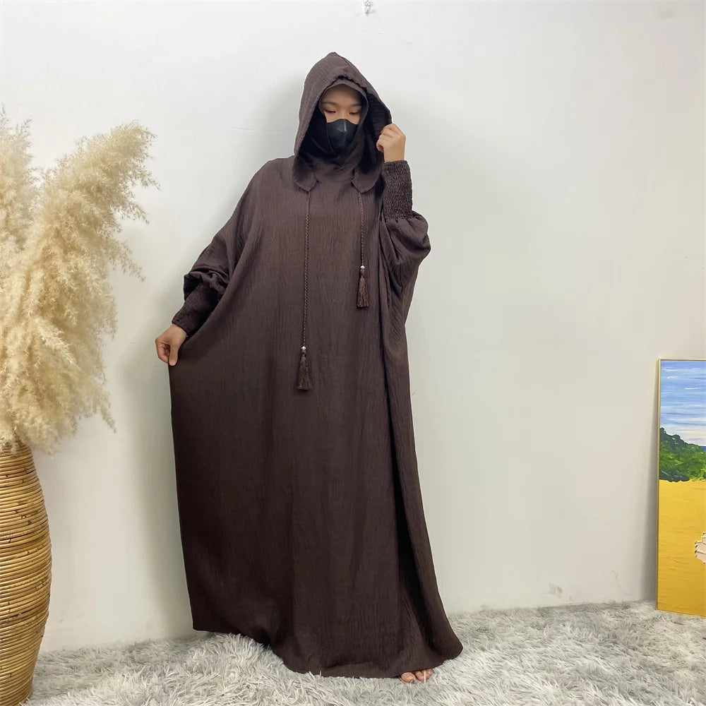Une femme portant une abaya marron foncé avec capuche et manches batwing se tient contre un mur blanc.