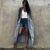 Eine Frau in einem hellblauen gemusterten Kimono-Überwurf über einem weißen Top und Jeansshorts steht vor einer Betonwand.