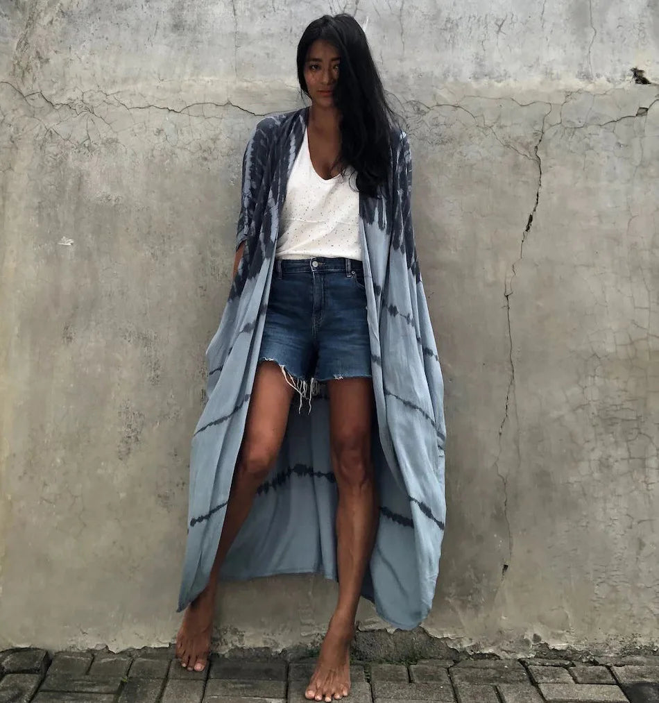 Eine Frau in einem hellblauen gemusterten Kimono-Überwurf über einem weißen Top und Jeansshorts steht vor einer Betonwand.
