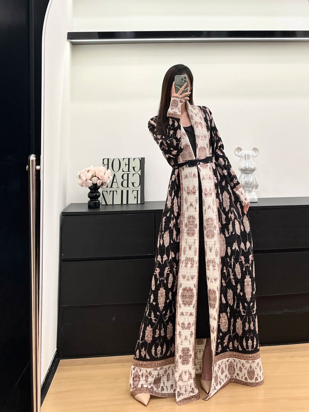 Une femme modélise un long trench à motifs noir et crème dans une pièce lumineuse.