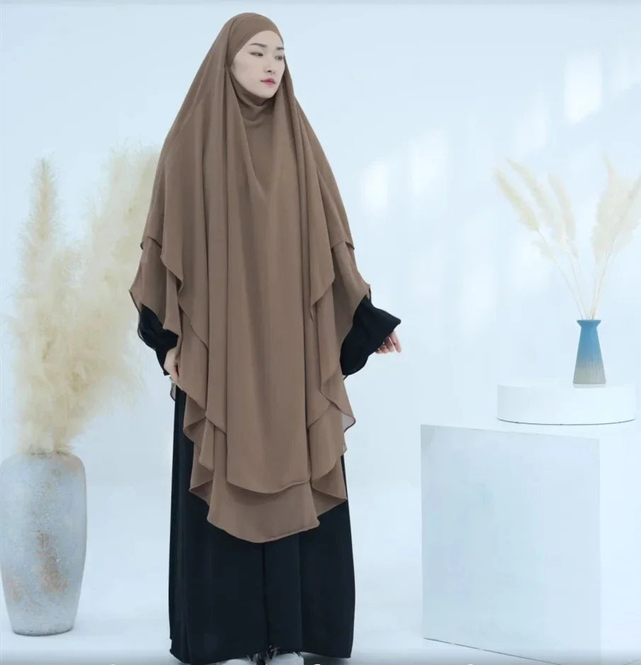 Eine Frau trägt einen beigen, zweilagigen Chiffon-Niqab und schwarze Kleidung vor weißem Hintergrund.