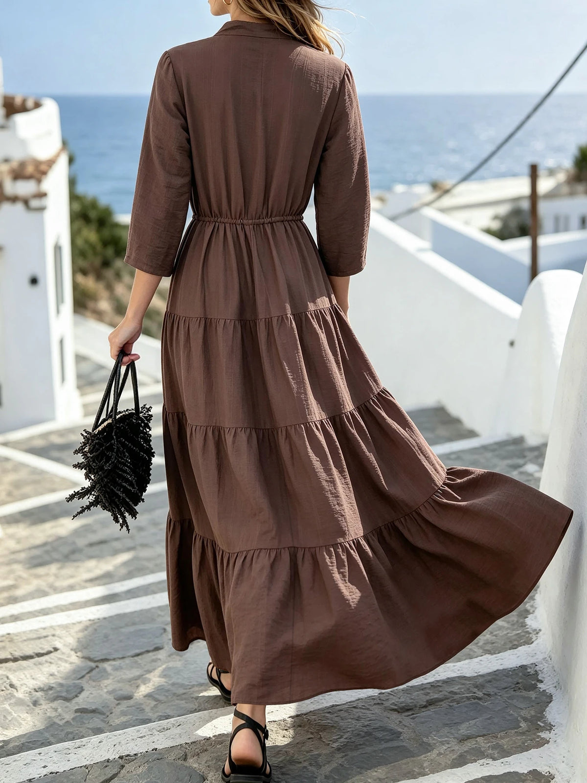 Eine Frau trägt ein langes, braunes Maxi-Kleid mit Zierzug und Rüschensaum, steht draußen.