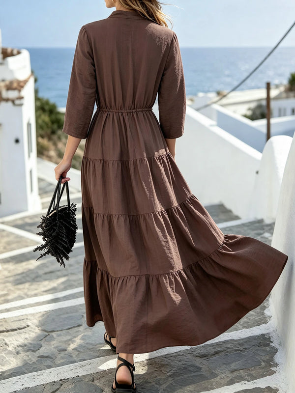A woman wearing a long, brown maxi dress with a drawstring waist and ruffle hem, standing outdoors.