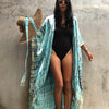 Eine Frau in einem hellblauen gemusterten Kimono-Überkleid über einem schwarzen Badeanzug steht vor einer Betonwand.