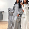 Una mujer modela una abaya de satin plata con corte batwing y un vestido blanco en una habitación luminosa.