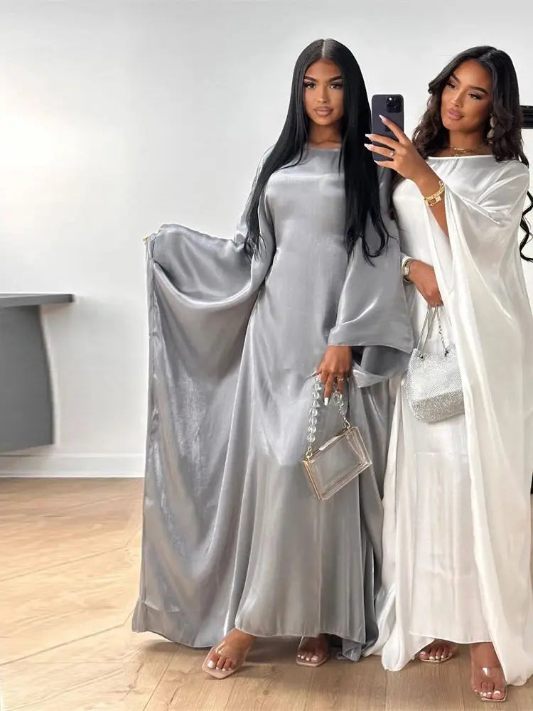 Una mujer modela una abaya de satin plata con corte batwing y un vestido blanco en una habitación luminosa.
