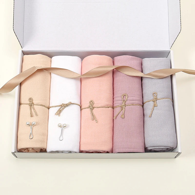 Une boîte contenant quatre foulards de hijab unis roulés en beige, blanc, rose et gris, attachés avec un ruban.