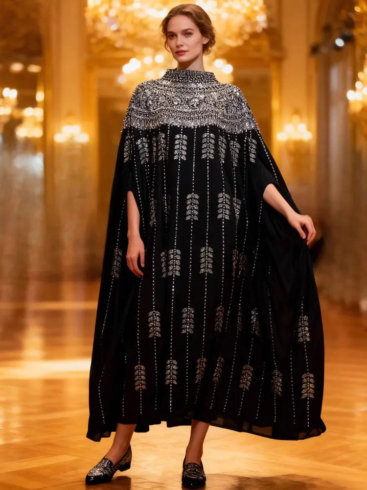 Model in black batwing robe dress heavily adorned with silver sequins and embroidery walking on a wooden runway.