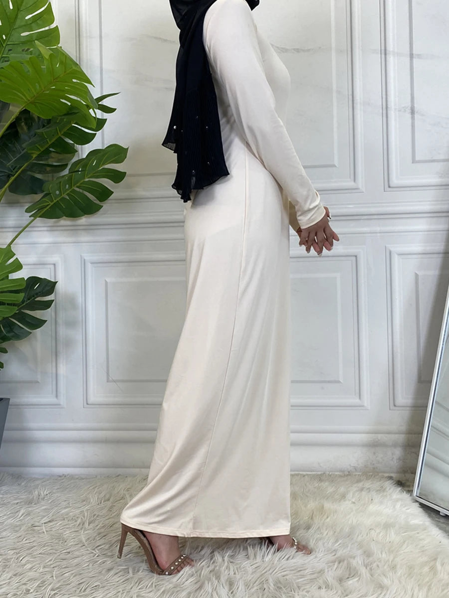 Eine Frau in einem eleganten cremefarbenen Maxi-Abaya-Kleid und einem schwarzen Hijab vor weiß verkleidetem Hintergrund