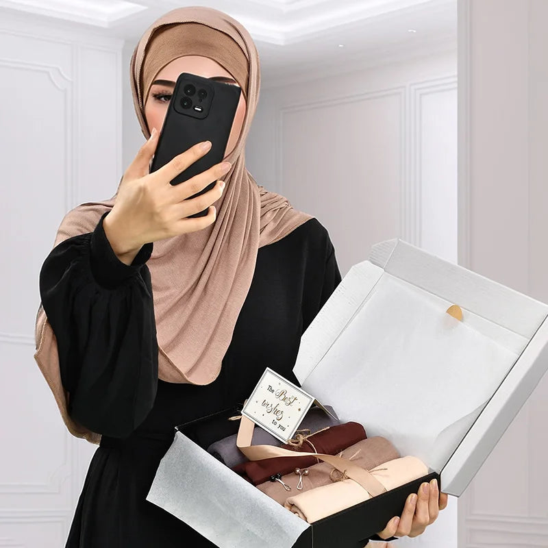 Une femme portant un hijab beige tient un smartphone noir en regardant une boîte cadeau blanche contenant un article en cuir 