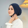 Eine Frau in einer dunkelblauen Innerhijab-Kappe und weißem Oberteil, posiert neben einer Leuchte.