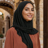 Une femme portant un foulard instant noir et un haut couleur rouille, dans un cadre architectural extérieur.