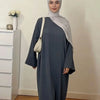 Une femme portant une abaya ample de couleur unie et un hijab gris clair se tient dans une pièce bien éclairée.