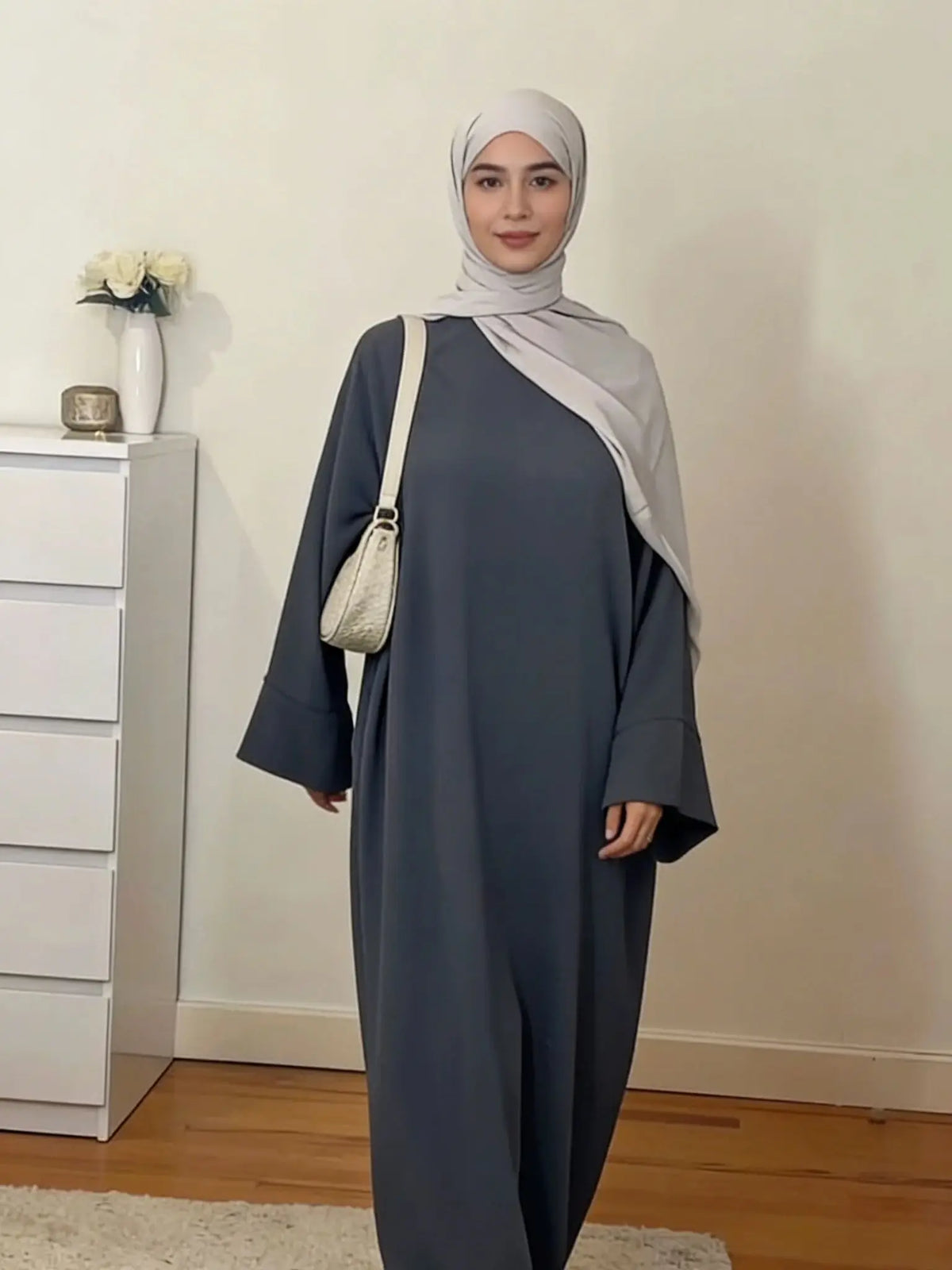 Une femme portant une abaya ample de couleur unie et un hijab gris clair se tient dans une pièce bien éclairée.