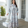 Une femme modélise une élégante abaya longue perlée de diamants avec des motifs floraux blancs et bleus, en extérieur.
