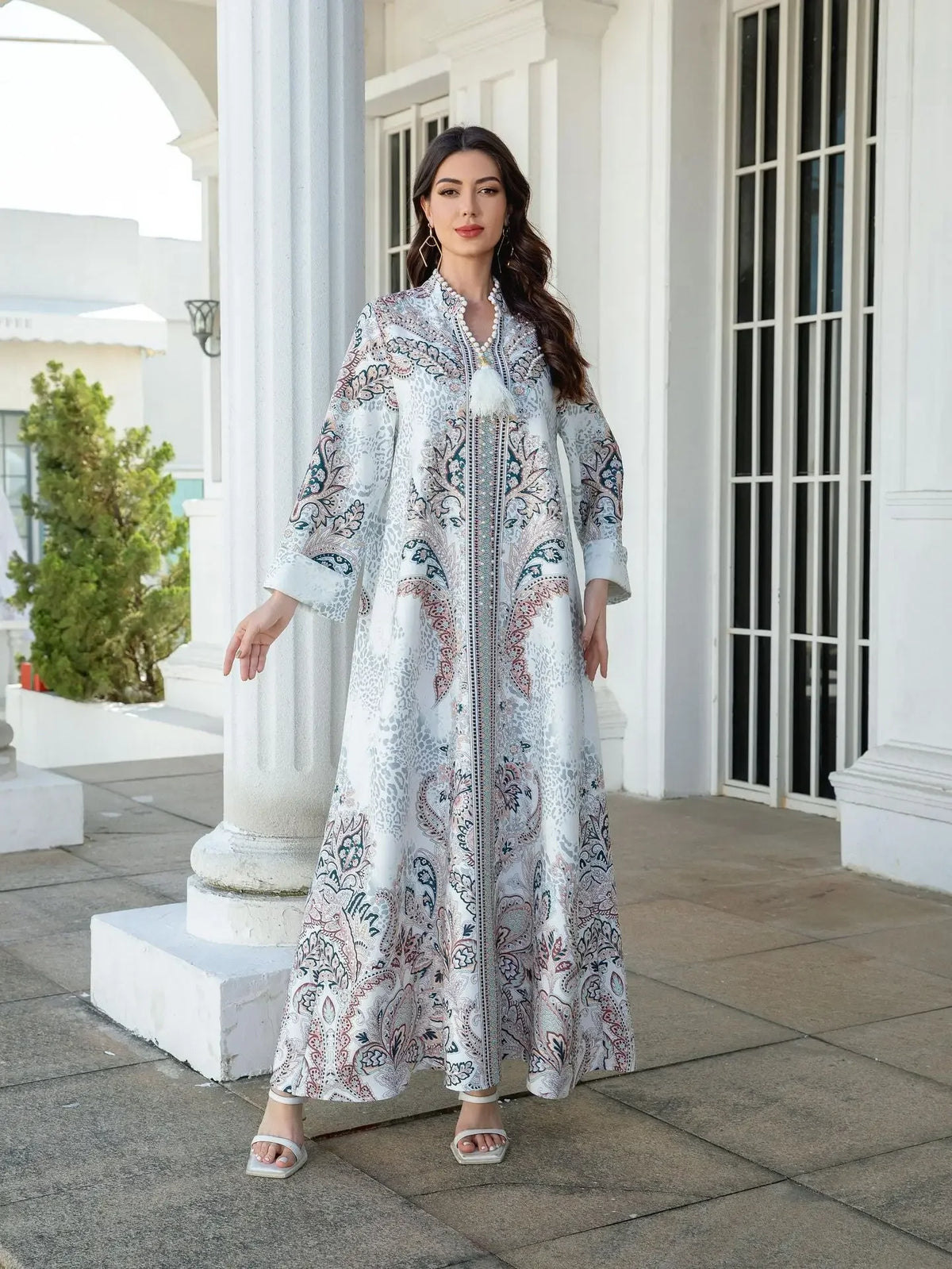 Une femme modélise une élégante abaya longue perlée de diamants avec des motifs floraux blancs et bleus, en extérieur.