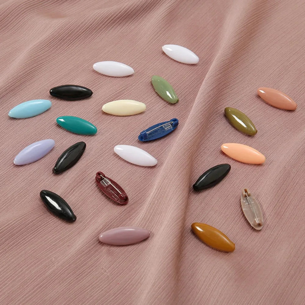 A collection of eight solid color brooch pins in various shades including white, green, blue, and peach, laid out on pink fab