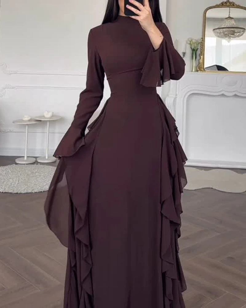 A woman wearing a dark plum long-sleeve maxi dress with ruffled details standing in a bright room.