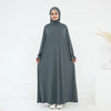 Una mujer vistiendo un abaya suelto y fluido gris oscuro con hijab, de pie contra un fondo blanco.