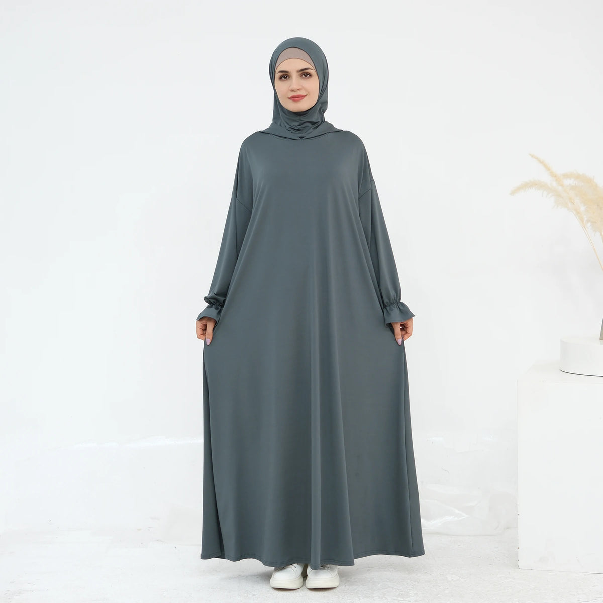 Una mujer vistiendo un abaya suelto y fluido gris oscuro con hijab, de pie contra un fondo blanco.