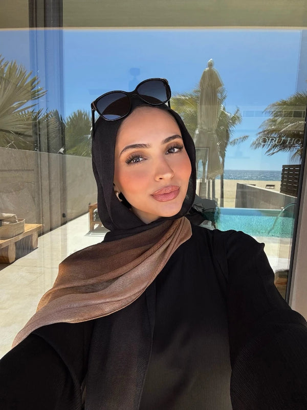 A woman wearing a black ombre gradient rayon cotton hijab scarf and sunglasses, posing in front of a glass window.